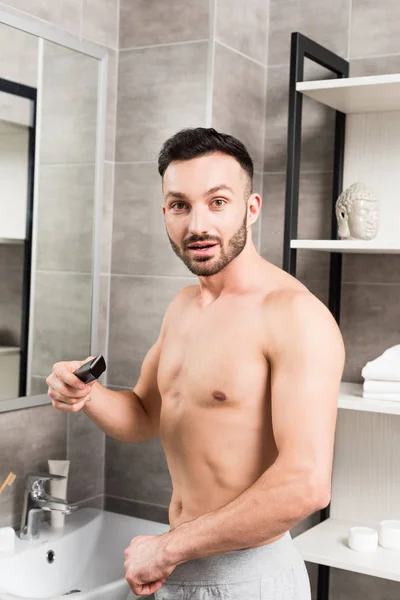 Sorprendido hombre sin camisa que sostiene trimmer mientras está de pie en el baño - foto de stock