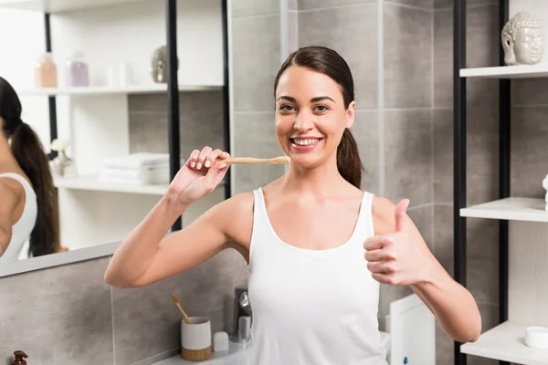 Cheerful woman showing thumb up while holding toothbrush in bathroom — Stock Photo