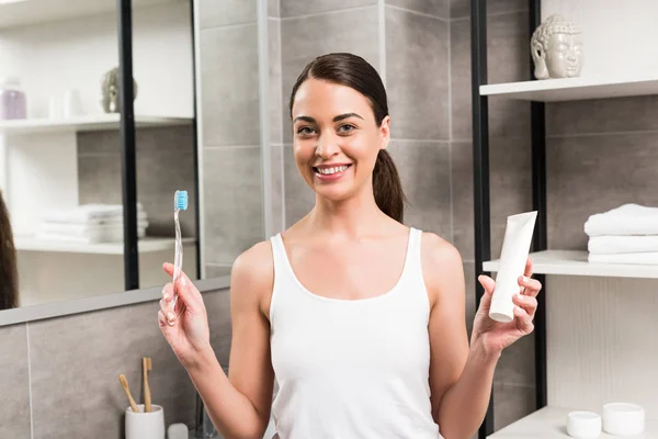 Alegre morena mujer sosteniendo cepillo de dientes y pasta de dientes en el baño - foto de stock