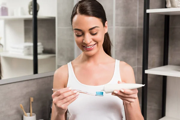 Cheerful brunette woman holding toothbrush while applying toothpaste in bathroom — Stock Photo