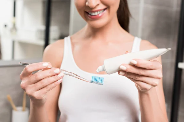 Selective focus of cheerful woman holding toothbrush while applying toothpaste in bathroom — Stock Photo