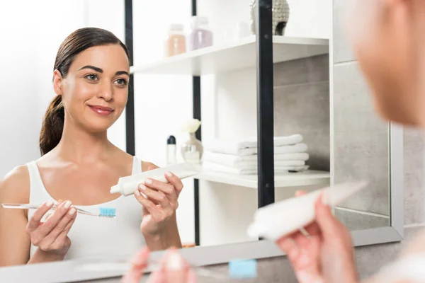 Foco seletivo de mulher morena alegre segurando escova de dentes e pasta de dentes enquanto olha para o espelho no banheiro — Fotografia de Stock