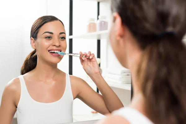 Selektiver Fokus der fröhlichen brünetten Frau beim Zähneputzen, während sie im Badezimmer in den Spiegel schaut — Stockfoto