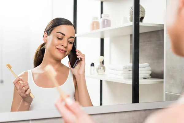 Foco seletivo de mulher morena feliz segurando escova de dentes enquanto conversa no smartphone no banheiro — Fotografia de Stock