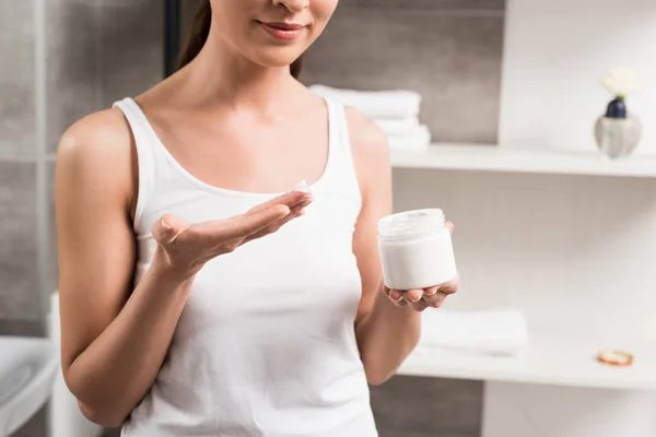 Vista recortada de la mujer alegre sosteniendo contenedor con crema facial en el baño - foto de stock