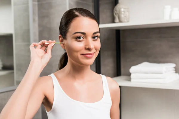 Atractiva mujer morena sonriendo mientras limpia el oído en el baño - foto de stock