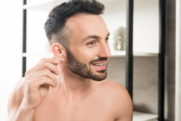 Cheerful shirtless man cleaning ear while looking at mirror in bathroom — Stock Photo