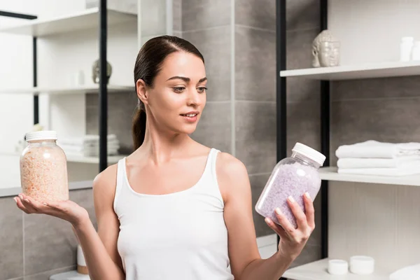 Atractiva mujer morena sosteniendo frascos de vidrio con sal marina en el baño - foto de stock