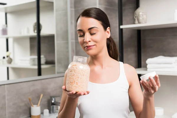 Attractive brunette woman smelling sea salt while holding glass jar in bathroom — Stock Photo