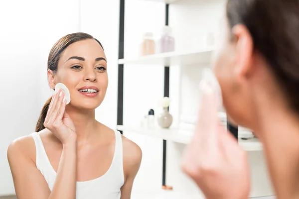 Selective focus of attractive brunette woman holding cotton pad near face while looking at mirror in bathroom — Stock Photo