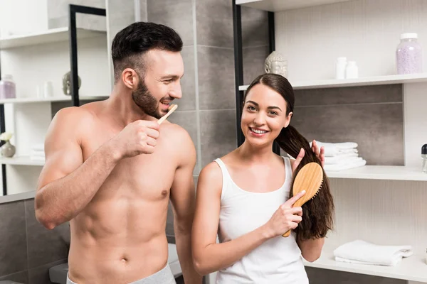 Homem alegre olhando para namorada atraente escovando o cabelo no banheiro — Fotografia de Stock