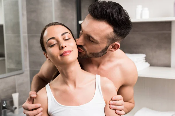 Handsome bearded man kissing cheek of happy brunette girlfriend in bathroom — Stock Photo
