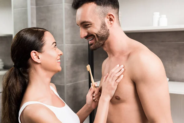 Alegre morena namorada segurando escova de dentes ao abraçar feliz namorado sem camisa no banheiro — Fotografia de Stock