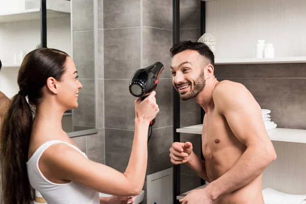 Mulher brincalhão segurando secador de cabelo enquanto de pé com namorado alegre no banheiro — Fotografia de Stock