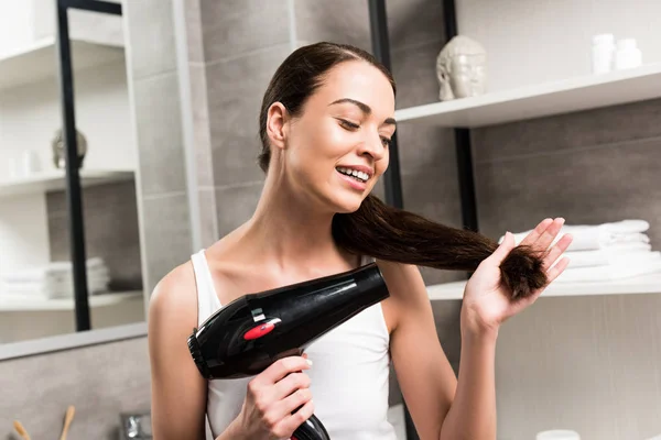 Happy brunette woman holding hair dryer while standing in bathroom — Stock Photo