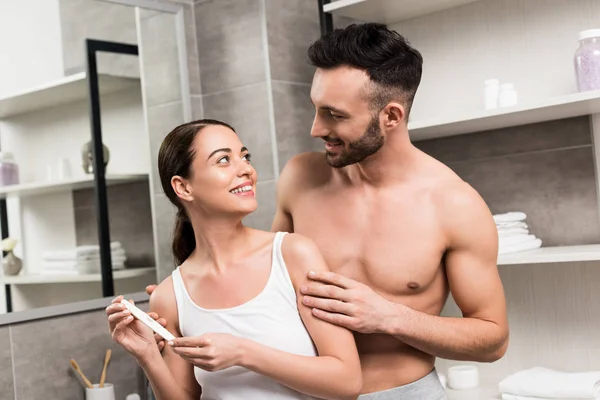 Feliz hombre sin camisa mirando alegre novia celebración prueba de embarazo en cuarto de baño - foto de stock