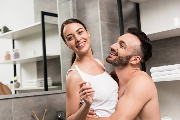 Excitado sin camisa hombre abrazando alegre novia celebración embarazo prueba en cuarto de baño - foto de stock