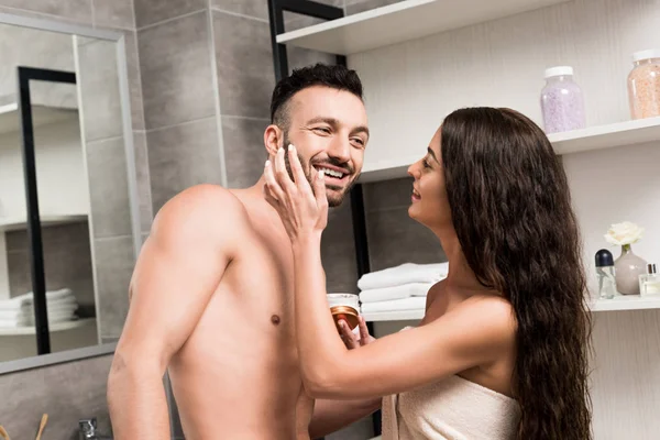 Happy woman applying cosmetic cream on cheek of happy bearded boyfriend — Stock Photo