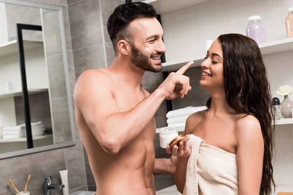 Alegre sin camisa novio aplicando crema facial en la nariz de morena novia - foto de stock