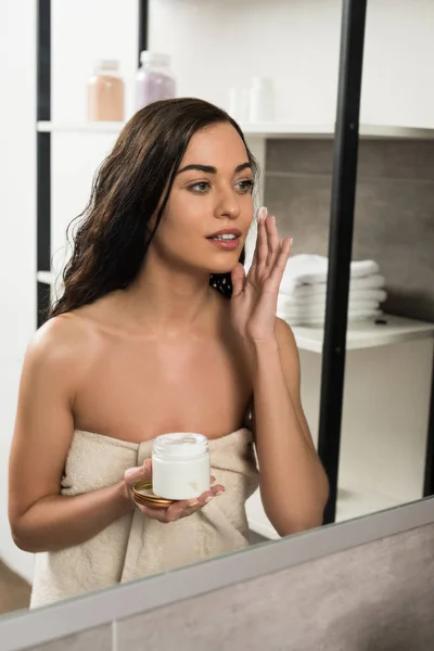 Привлекательная женщина наносит крем для лица на щеку, глядя в зеркало в ванной комнате — стоковое фото