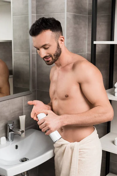 Hombre sin camisa guapo sosteniendo lata con espuma de afeitar en el baño - foto de stock