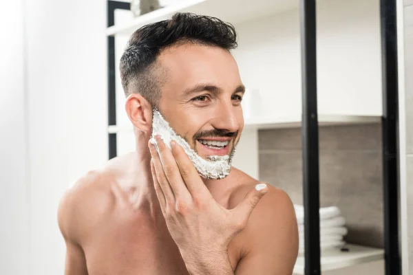 Uomo barbuto allegro applicare schiuma da barba sul viso in bagno — Foto stock