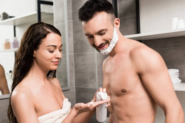 Hombre barbudo guapo con espuma de afeitar en la cara mirando a la mano de novia atractiva en el baño - foto de stock