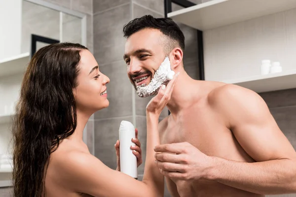 Приваблива дівчина наносить піну для гоління на обличчя красивого бородатого чоловіка у ванній — стокове фото