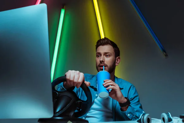 Handsome and good-looking man drinking soda and playing video game with steering wheel — Stock Photo