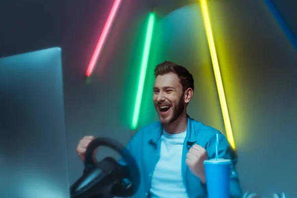Избирательный фокус красивого и улыбающегося мужчины, играющего в видеоигру с рулем — стоковое фото