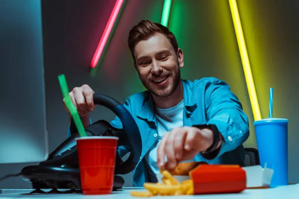 Selective focus of handsome and smiling man holding steering wheel and eating french fries — Stock Photo