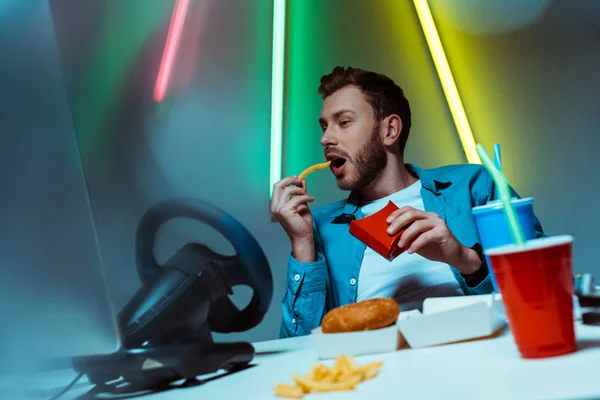 Good-looking and handsome man eating french fries and looking at computer monitor — Stock Photo