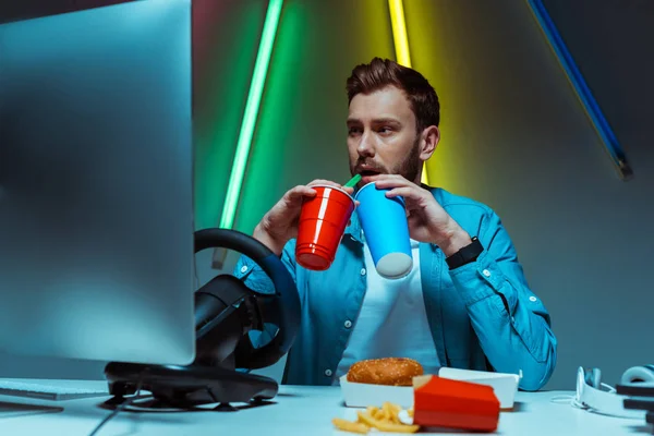 Homem bonito e bonito bebendo de copos de plástico e papel e olhando para o monitor — Fotografia de Stock