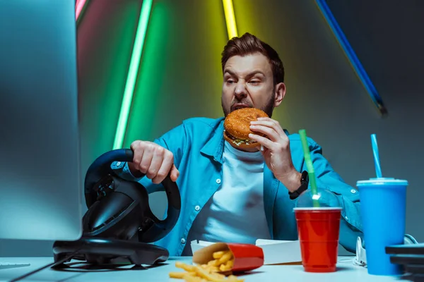 Handsome and good-looking cyber sportsman eating burger and playing computer game — Stock Photo