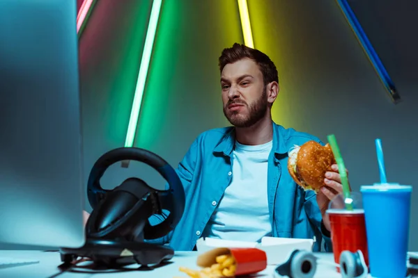 Selektiver Fokus eines gutaussehenden Mannes, der Burger isst und auf den Computermonitor blickt — Stockfoto