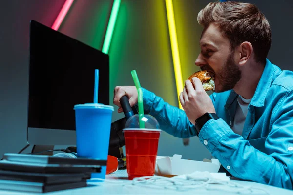 Selective focus of handsome and good-looking cyber sportsman eating tasty burger and playing video game — Stock Photo