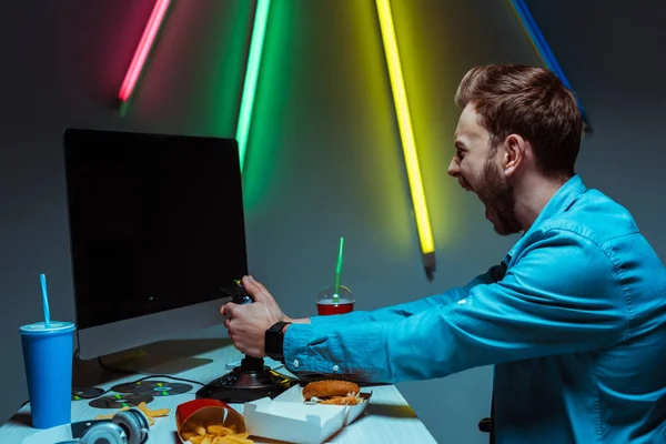 Bonito e irritado homem gritando, segurando joystick e olhando para monitor de computador — Fotografia de Stock