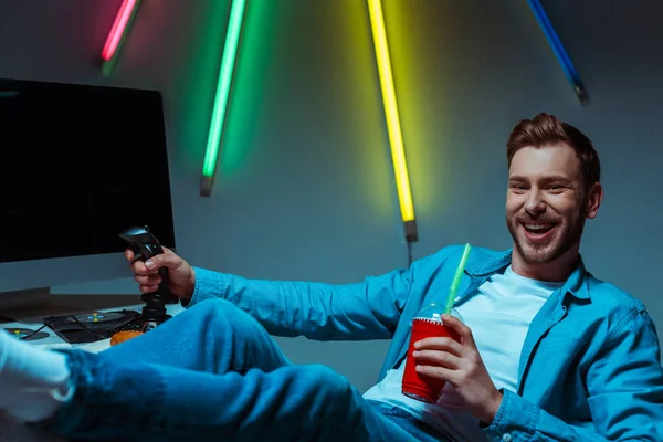 Selective focus of smiling cyber sportsman holding joystick, plastic cup and playing video game — Stock Photo