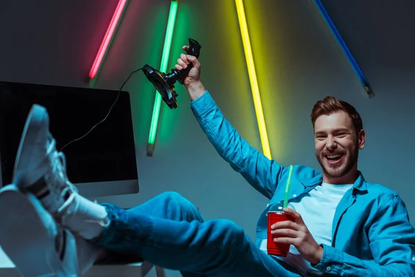 Foco seletivo de sorrindo desportista cibernético segurando joystick e copo de plástico com bebida — Fotografia de Stock