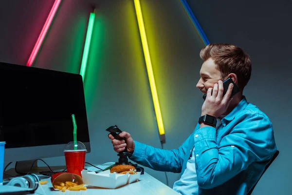 Вид сбоку улыбающегося и красивого киберчеловека, держащего джойстик и говорящего по смартфону — стоковое фото