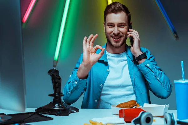 Sonriente y guapo hombre hablando en el teléfono inteligente y mostrando buen gesto - foto de stock