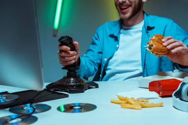 Selective focus of man holding tasty burger and playing video game with joystick — Stock Photo