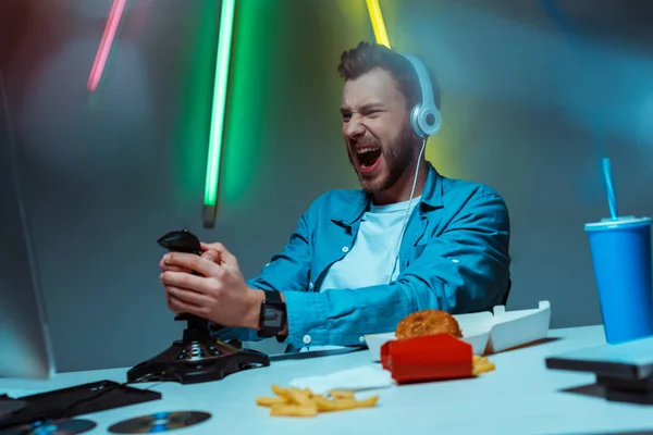 Enfoque selectivo de cyber sportsman enojado en auriculares jugando videojuego con joystick - foto de stock