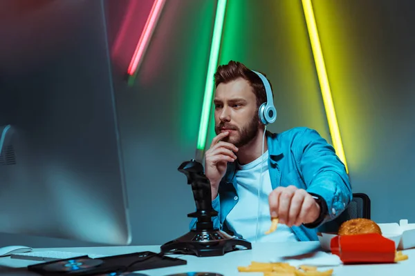 Selektiver Fokus eines gut aussehenden Cyber-Mannes mit Kopfhörern, der auf den Computermonitor schaut und Pommes frites isst — Stockfoto