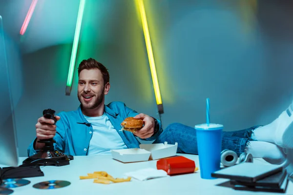 Apuesto deportista cibernético jugando videojuego con joystick y la celebración de hamburguesa - foto de stock