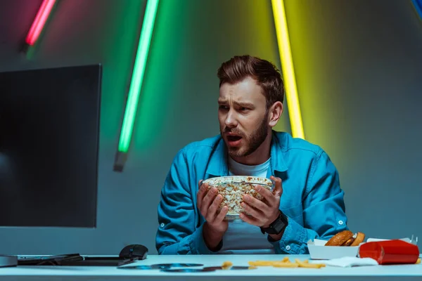 Handsome and shocked man holding bowl with popcorn and looking at computer monitor — Stock Photo