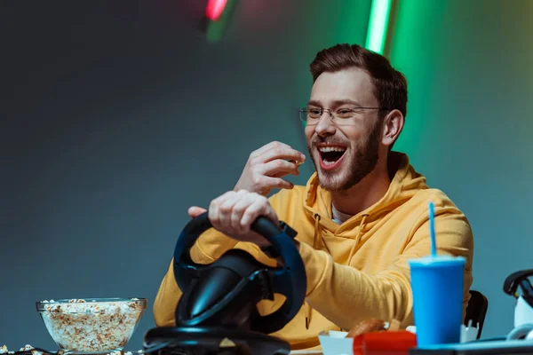 Uomo sorridente e di bell'aspetto in occhiali che gioca con il volante e mangia popcorn — Foto stock