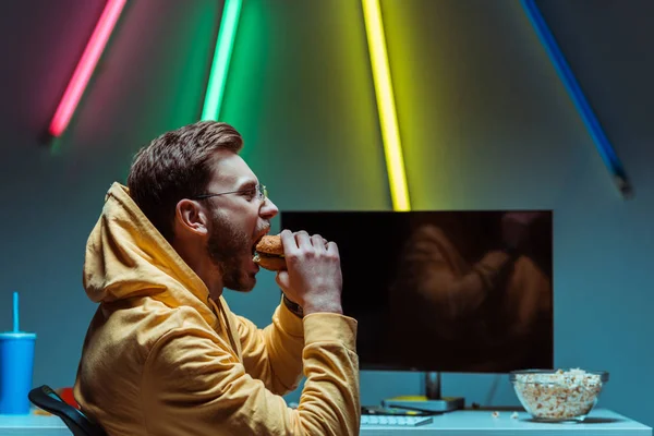 Side view of handsome and young adult man eating tasty burger and looking away — Stock Photo