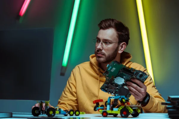 Homem bonito e bonito em óculos segurando placa de circuito de computador e olhando para monitor de computador — Fotografia de Stock