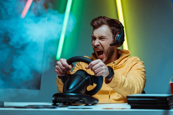Angry and handsome man in headphones playing video game with steering wheel — Stock Photo